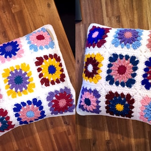 Könnte beinhalten: Ein weißes gehäkeltes Kissen mit einem bunten Blumenmuster. Das Kissen besteht aus neun Quadraten, jedes mit einem anderen farbigen Blumendesign. Die Blumen sind in den Farben Rosa, Blau, Gelb, Rot und Lila.