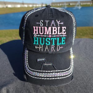 May include: Black baseball cap with the embroidered text "STAY HUMBLE HUSTLE HARD" in pink and teal. The cap has a distressed look with decorative stitching and a curved brim. The cap is made of a durable fabric.