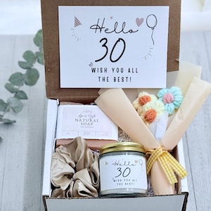 May include: A gift box containing a bar of natural soap, a gold-colored satin scrunchie, a small candle with a label reading Hello 30 WISH YOU ALL THE BEST, and a small bouquet of crocheted flowers wrapped in paper with a yellow gingham ribbon.