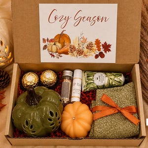 May include: A cardboard gift box containing a green ceramic pumpkin, an orange pumpkin candle, a tube of hand cream, a lip balm, a small glass bottle of matches, two gold-wrapped chocolates, and a green knit item tied with an orange ribbon.