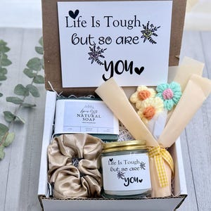May include: A gift box containing a bar of natural soap, a gold scrunchie, a candle, and a small bouquet of crocheted flowers. The candle and card read "Life Is Tough but so are you."