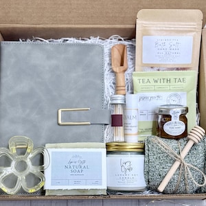 May include: A gift box filled with self-care items. Includes a gray journal, soap, candle, bath salts, tea, honey, a wooden honey dipper, and a hair clip. The box is lined with tissue paper and tied with twine.