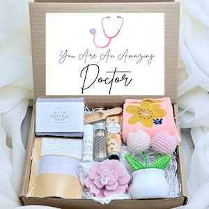 May include: A cardboard gift box containing a bar of natural soap, a small wooden scoop, a lip balm, a glass vial, a tube of honey hand cream, a pink flower-patterned cloth, a pink succulent-shaped candle, and two crocheted tulips in a white pot. The lid features a pink stethoscope graphic and the text: You Are An Amazing Doctor.