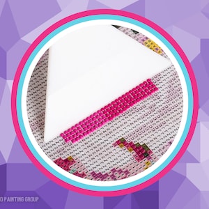 May include: A close-up of a diamond painting in progress. A white tray with pink diamonds is placed on top of a diamond painting canvas with a pink, blue, and white circle border. The canvas has a grid pattern with numbers and letters.