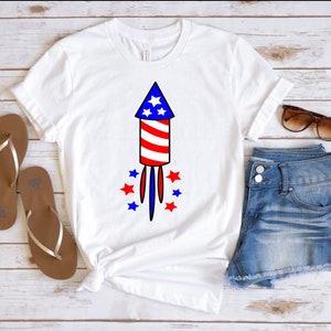 May include: White t-shirt with a red, white, and blue firework graphic. The firework has a star on top and is surrounded by smaller stars. The shirt is on a wooden surface with a pair of brown sandals and blue denim shorts.
