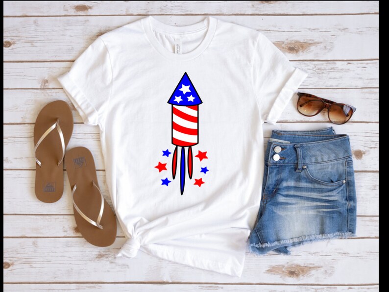 May include: White t-shirt with a red, white, and blue firework graphic. The firework has a star on top and is surrounded by smaller stars. The shirt is on a wooden surface with a pair of brown sandals and blue denim shorts.