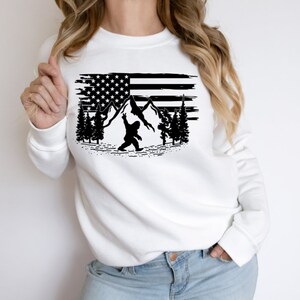 May include: White sweatshirt with a black graphic of a Bigfoot holding a flag with mountains and trees in the background.