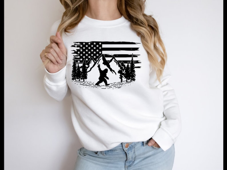 May include: White sweatshirt with a black graphic of a Bigfoot holding a flag with mountains and trees in the background.