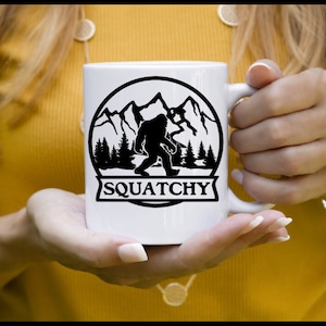 May include: White ceramic mug with a black silhouette of Bigfoot in a mountain landscape. The text "SQUATCHY" is printed below the image.
