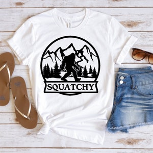 May include: White t-shirt with a black graphic of a Bigfoot in a mountain scene. The graphic is inside a circle with the text "SQUATCHY" below it.