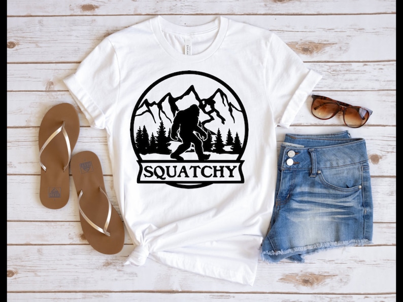May include: White t-shirt with a black graphic of a Bigfoot in a mountain scene. The graphic is inside a circle with the text "SQUATCHY" below it.