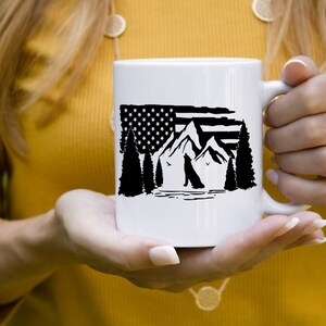 Puede incluir: Taza de cer&aacute;mica blanca con un dise&ntilde;o en blanco y negro de monta&ntilde;as, &aacute;rboles y la silueta de un perro, incorporando una bandera estadounidense. La taza tiene un asa curva y la sostiene una persona.