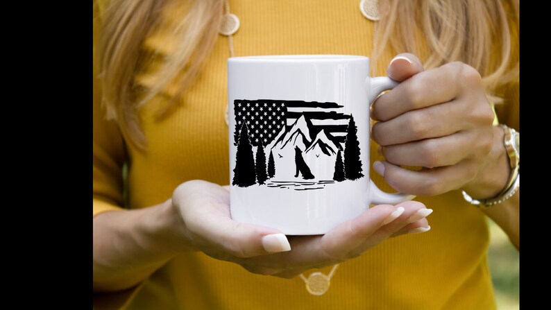 Puede incluir: Taza de cer&aacute;mica blanca con un dise&ntilde;o en blanco y negro de monta&ntilde;as, &aacute;rboles y la silueta de un perro, incorporando una bandera estadounidense. La taza tiene un asa curva y la sostiene una persona.