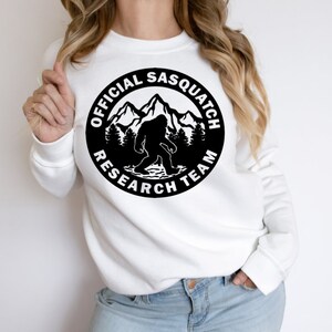 May include: White sweatshirt with a black circle graphic featuring a silhouette of Bigfoot walking in front of mountains and trees. The text around the graphic reads "Official Sasquatch Research Team."