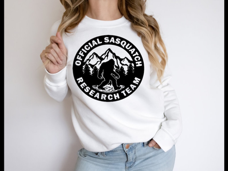 May include: White sweatshirt with a black circle graphic featuring a silhouette of Bigfoot walking in front of mountains and trees. The text around the graphic reads "Official Sasquatch Research Team."