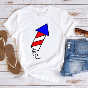 May include: White t-shirt with a red, white, and blue firework graphic. The firework is shaped like an arrow pointing up. The shirt is paired with denim shorts and brown sandals.