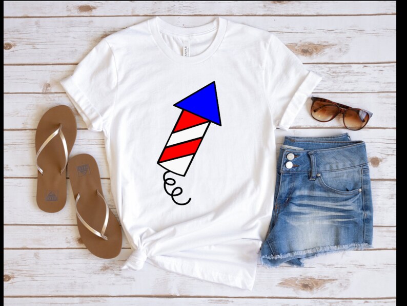May include: White t-shirt with a red, white, and blue firework graphic. The firework is shaped like an arrow pointing up. The shirt is paired with denim shorts and brown sandals.