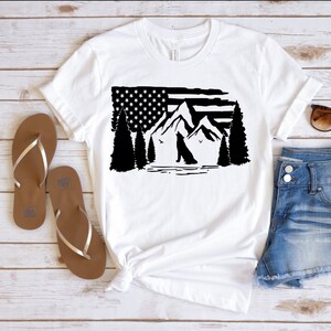 Puede incluir: Camiseta blanca con un gr&aacute;fico en blanco y negro de la bandera estadounidense, monta&ntilde;as, &aacute;rboles y un lobo. La camiseta se combina con sandalias marrones, pantalones cortos de mezclilla y gafas de sol sobre una superficie de madera clara.