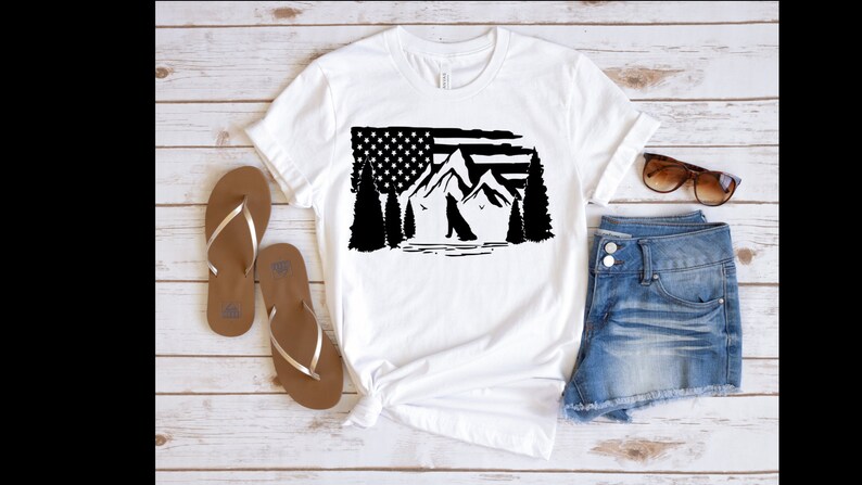 Puede incluir: Camiseta blanca con un gr&aacute;fico en blanco y negro de la bandera estadounidense, monta&ntilde;as, &aacute;rboles y un lobo. La camiseta se combina con sandalias marrones, pantalones cortos de mezclilla y gafas de sol sobre una superficie de madera clara.