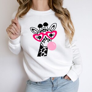 Puede incluir: Sudadera blanca con una jirafa en blanco y negro que lleva gafas de coraz&oacute;n rosa y sopla una burbuja rosa.