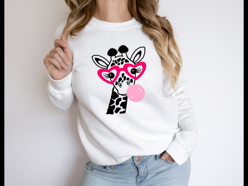Puede incluir: Sudadera blanca con una jirafa en blanco y negro que lleva gafas de coraz&oacute;n rosa y sopla una burbuja rosa.