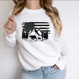 Puede incluir: Sudadera blanca con un dise&ntilde;o gr&aacute;fico negro de la bandera estadounidense, monta&ntilde;as, &aacute;rboles y osos. El dise&ntilde;o tiene un aspecto desgastado. La sudadera es de cuello redondo y manga larga.
