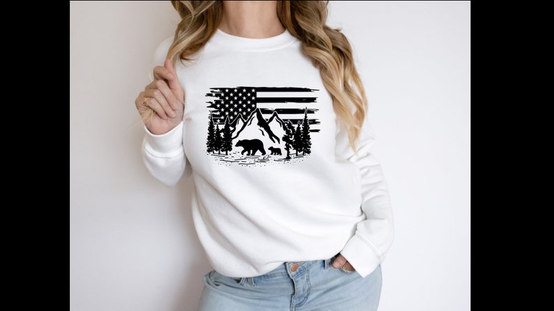 Puede incluir: Sudadera blanca con un dise&ntilde;o gr&aacute;fico negro de la bandera estadounidense, monta&ntilde;as, &aacute;rboles y osos. El dise&ntilde;o tiene un aspecto desgastado. La sudadera es de cuello redondo y manga larga.