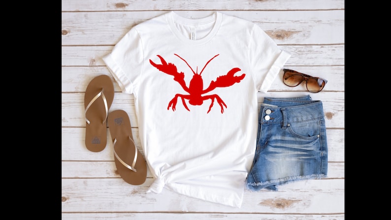 K&ouml;nnte beinhalten: Wei&szlig;es T-Shirt mit einem roten Hummer-Silhouetten-Motiv. Das Shirt wird mit Jeans-Shorts und braunen Flip-Flops kombiniert.