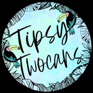 May include: A circular design with a watercolor background in shades of blue and green. The text "Tipsy Two Cans" is written in black cursive font. Two toucans with pink beaks and black bodies are on either side of the text.