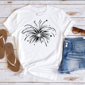May include: White t-shirt with a black firework graphic. The shirt is styled with denim shorts and brown sandals.