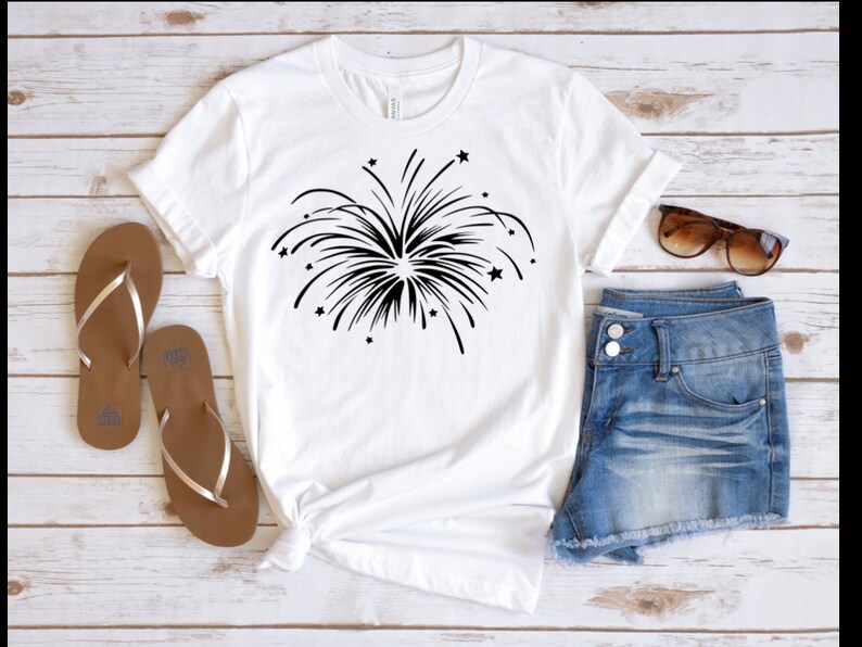 May include: White t-shirt with a black firework graphic. The shirt is styled with denim shorts and brown sandals.