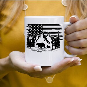 Puede incluir: Taza de cer&aacute;mica blanca con un dise&ntilde;o en blanco y negro de la bandera estadounidense, monta&ntilde;as, &aacute;rboles y osos. La taza tiene un asa. El dise&ntilde;o es una ilustraci&oacute;n patri&oacute;tica y con tem&aacute;tica de la naturaleza.