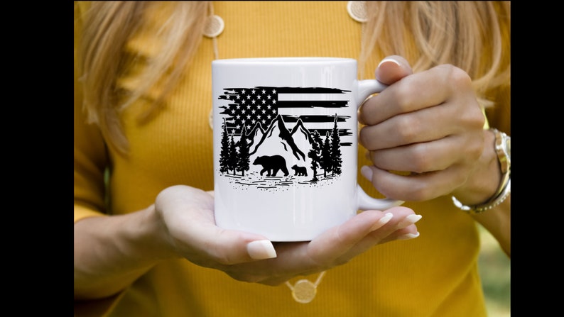 Puede incluir: Taza de cer&aacute;mica blanca con un dise&ntilde;o en blanco y negro de la bandera estadounidense, monta&ntilde;as, &aacute;rboles y osos. La taza tiene un asa. El dise&ntilde;o es una ilustraci&oacute;n patri&oacute;tica y con tem&aacute;tica de la naturaleza.