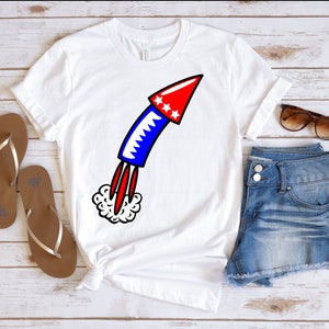 May include: White t-shirt with a red, white, and blue firework graphic. The firework is a cartoon style rocket with a red top and blue body. The rocket is launching with a white smoke trail.