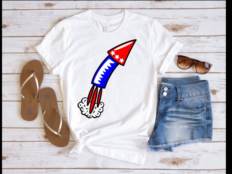 May include: White t-shirt with a red, white, and blue firework graphic. The firework is a cartoon style rocket with a red top and blue body. The rocket is launching with a white smoke trail.