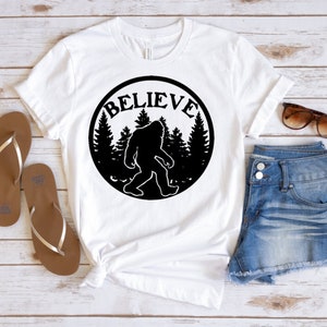 May include: White t-shirt with a black circle graphic featuring a silhouette of Bigfoot walking through a forest with the word "BELIEVE" written around the outside of the circle.