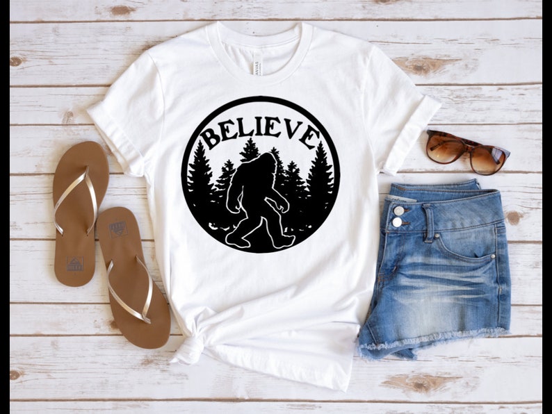 May include: White t-shirt with a black circle graphic featuring a silhouette of Bigfoot walking through a forest with the word "BELIEVE" written around the outside of the circle.
