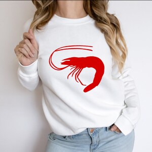 K&ouml;nnte beinhalten: Wei&szlig;er Pullover mit einem roten Garnelen-Motiv.