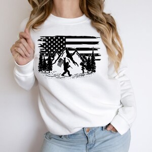 May include: White sweatshirt with a black graphic of a distressed American flag with mountains and a figure holding a flag in front of the flag.