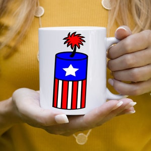 May include: White ceramic mug with a red, white, and blue firework design. The firework is shaped like a cylinder with a star on the blue section and red and white stripes on the bottom.