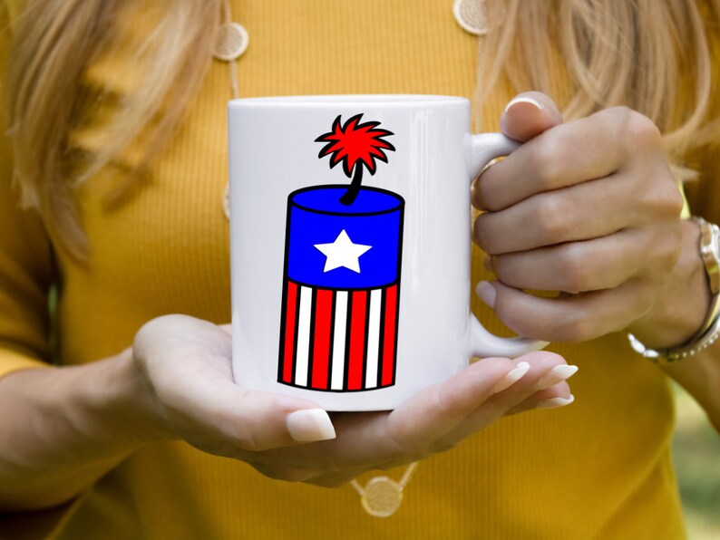 May include: White ceramic mug with a red, white, and blue firework design. The firework is shaped like a cylinder with a star on the blue section and red and white stripes on the bottom.