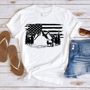 Puede incluir: Camiseta blanca con un gr&aacute;fico en blanco y negro de la bandera estadounidense, monta&ntilde;as, &aacute;rboles y un ciervo. La camiseta se combina con pantalones cortos de mezclilla y sandalias marrones. Gafas de sol sobre los pantalones cortos.