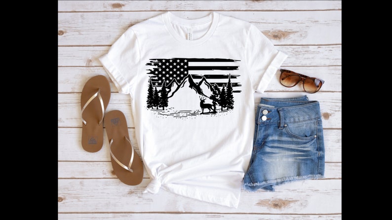 Puede incluir: Camiseta blanca con un gr&aacute;fico en blanco y negro de la bandera estadounidense, monta&ntilde;as, &aacute;rboles y un ciervo. La camiseta se combina con pantalones cortos de mezclilla y sandalias marrones. Gafas de sol sobre los pantalones cortos.