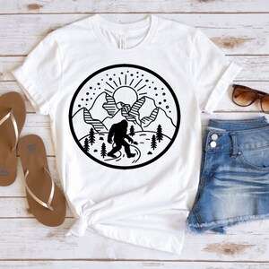 May include: White t-shirt with a black and white graphic design of a Bigfoot standing in front of mountains with a sun and stars in the sky.