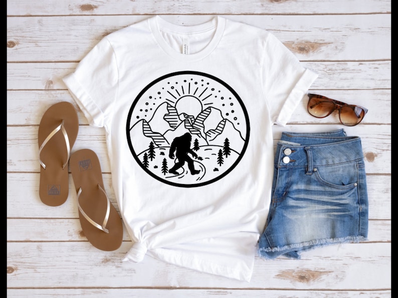 May include: White t-shirt with a black and white graphic design of a Bigfoot standing in front of mountains with a sun and stars in the sky.