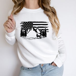 Puede incluir: Sudadera blanca con un gr&aacute;fico en blanco y negro de la bandera estadounidense, monta&ntilde;as, &aacute;rboles y un ciervo. El dise&ntilde;o tiene un efecto desgastado. La sudadera es de estilo casual y c&oacute;modo.