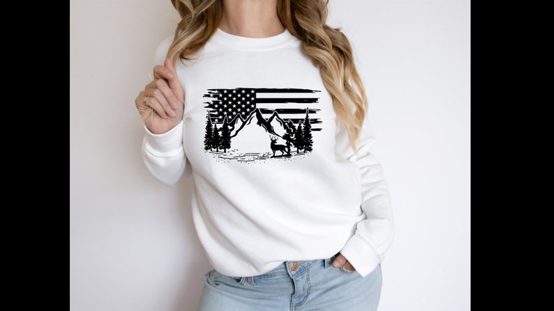 Puede incluir: Sudadera blanca con un gr&aacute;fico en blanco y negro de la bandera estadounidense, monta&ntilde;as, &aacute;rboles y un ciervo. El dise&ntilde;o tiene un efecto desgastado. La sudadera es de estilo casual y c&oacute;modo.