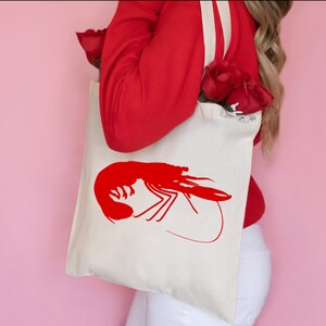 K&ouml;nnte beinhalten: Eine wei&szlig;e Einkaufstasche mit einem roten Garnelen-Silhouetten-Design. Die Tasche hat lange Henkel und wird von einer Person getragen, die einen roten Pullover und eine wei&szlig;e Hose tr&auml;gt.