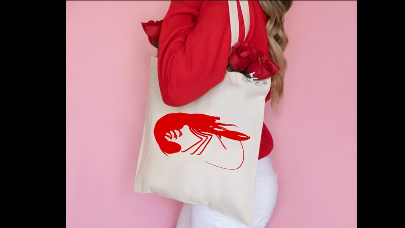 K&ouml;nnte beinhalten: Eine wei&szlig;e Einkaufstasche mit einem roten Garnelen-Silhouetten-Design. Die Tasche hat lange Henkel und wird von einer Person getragen, die einen roten Pullover und eine wei&szlig;e Hose tr&auml;gt.