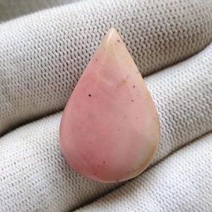 May include: A teardrop-shaped pink opal gemstone with a polished surface. The stone has a gradient of colors, transitioning from a pale beige at the top to a vibrant pink at the base. Small dark inclusions are visible on the surface.